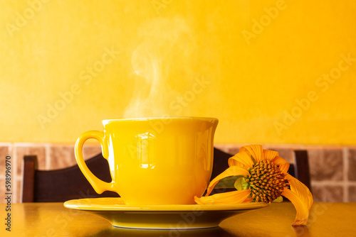 Uma xícara amarela cheia de café quente, com pires e um girassol murcho, sobre uma mesa de madeira com uma parede amarela ao fundo. photo