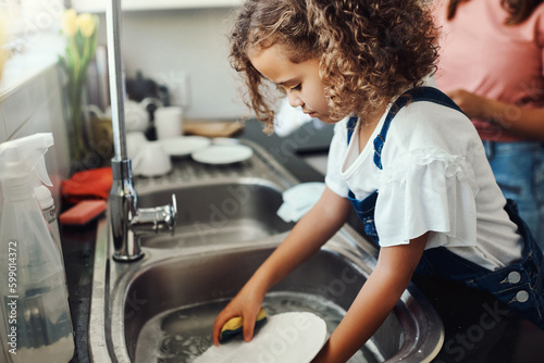 Moms got me doing the dishes. an adorable young girl standing and washing the dishes in the kitchen sink at home.