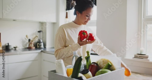 Young woman unpacking a grocery delivery box while standing at a table in her kitchen