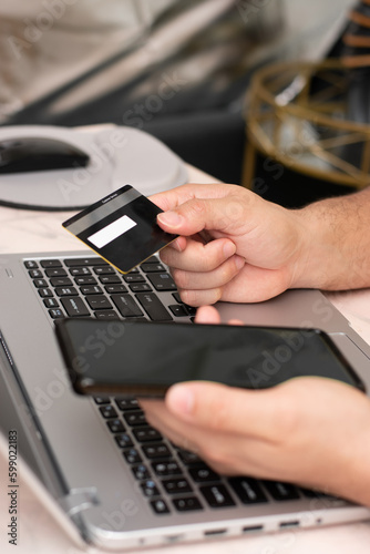 Vertical view of man buying with a black credit card and his phone front the laptop, online pay and shopping.
