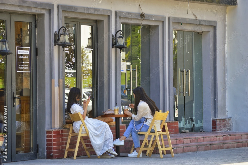 Obraz premium Two girls have breakfast in a summer street cafe in front of large mirrored cafe windows. Photos without faces and brands.