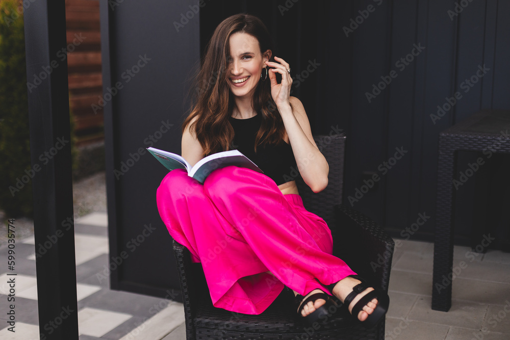 Young beautiful girl reading book while she is smiling and sitting on the armchair in her house outside. Happy woman have a rest and wear pink trousers and black top, read novel book, fix hair. Young beautiful girl reading book while she is smiling and sitting on the armchair in her house outside. Happy woman have a rest and wear pink trousers and black top, read novel book, fix hair.