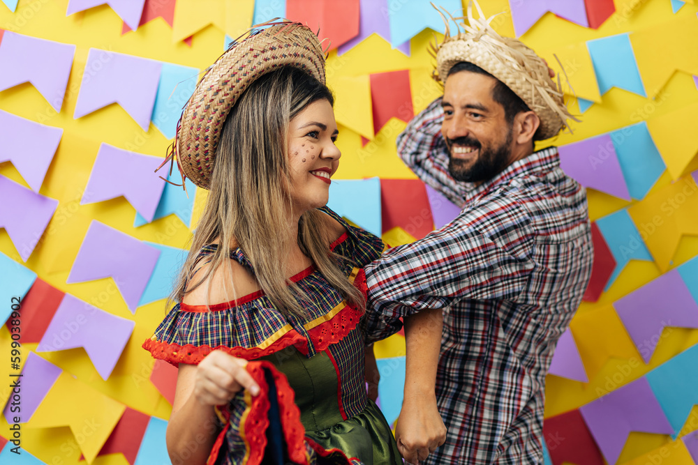 Brazilian couple wearing traditional clothes for Festa Junina - June festival - dancing over colorful background with pennants