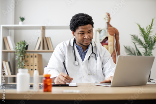 Focused african american man with stethoscope typing on laptop and making notes while sitting at writing desk with pill bottles on foreground. Efficient physician analyzing data via net in clinic.