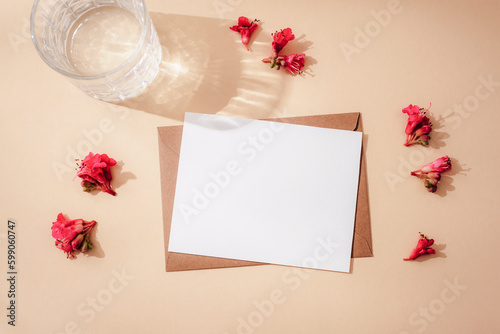 Envelope with blank card, red horse chestnut flowers and glass of water with harsh shadowson neutral beige background. Greeting card. Top view, flat lay, mockup photo