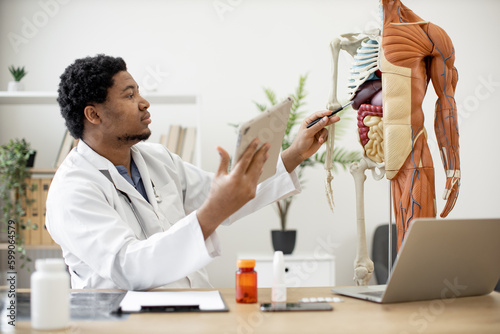 Fotografering Bearded african american male in lab coat marking liver on human anatomy model while sitting near desk with digital devices in workplace