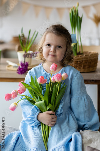 Cute little girl, baby daughter in a blue dress prepare pink tulip flowers to her happy mom. Celebration of Mother's or Women's day, birthday or anniversary. Cozy home atmosphere, kitchen. Springtime