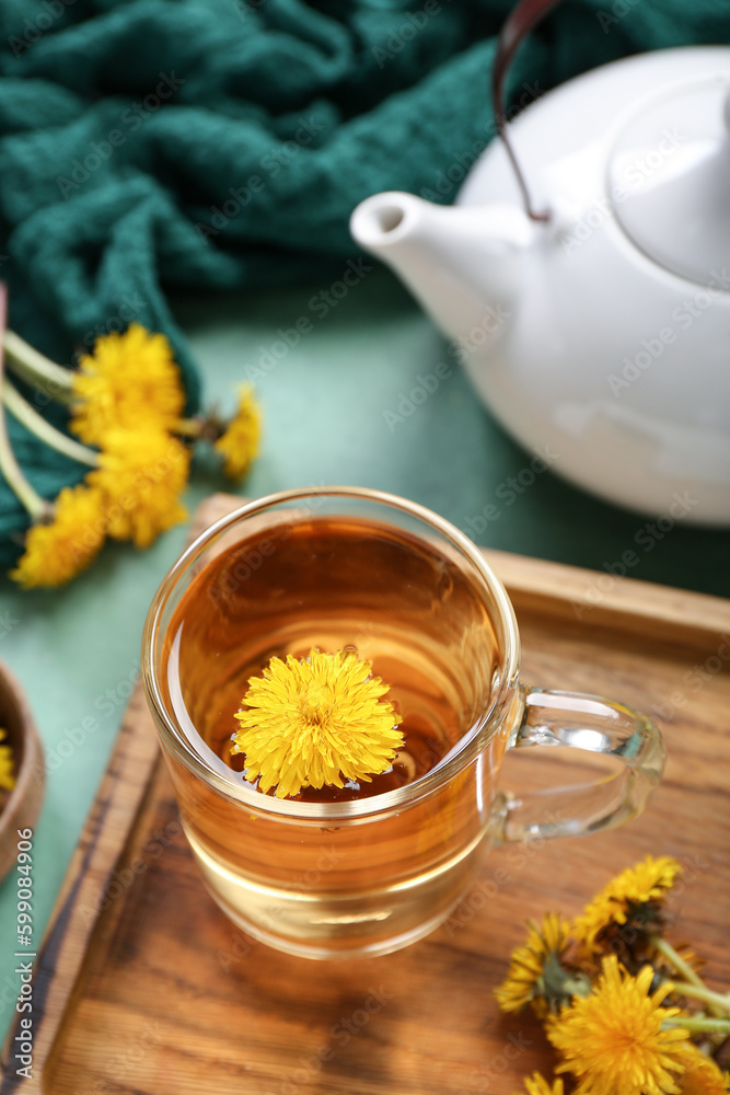 Board with glass cup of healthy dandelion tea and teapot on green background Board with glass cup of healthy dandelion tea and teapot on green background