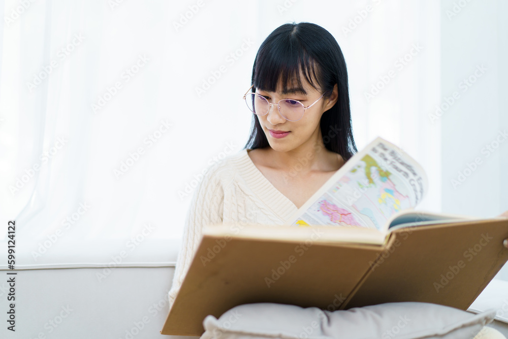 Happy - cheerful Asian young woman with eye glasses sitting on the sofa in living room and reading a book alone.
