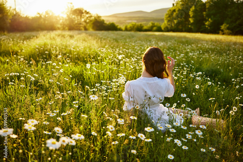 a red-haired woman in a light dress sits in a chamomile field at sunset and admires the passing day