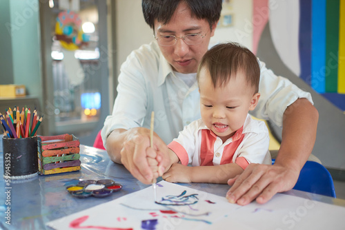 Cute smiling little Asian toddler baby boy child painting with brush and watercolors, Businessman father painting with son after working time, Creative play for toddlers, Soft and selective focus photo
