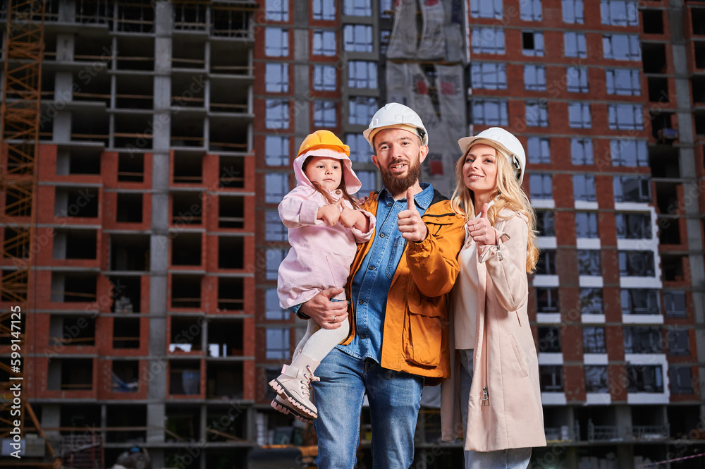Parents and daughter showing approval gesture and smiling while standing outdoors against apartment building under construction. Happy family homeowners giving thumbs up at construction site.