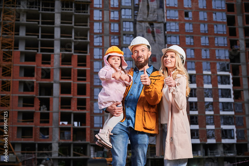 Parents and daughter showing approval gesture and smiling while standing outdoors against apartment building under construction. Happy family homeowners giving thumbs up at construction site. © anatoliy_gleb