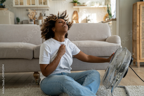 Summer heat. Young African American woman cooling down by ventilator at home, feeling unwell with high temperature during hot weather, sitting on floor in front of electric fan during extreme heatwave photo