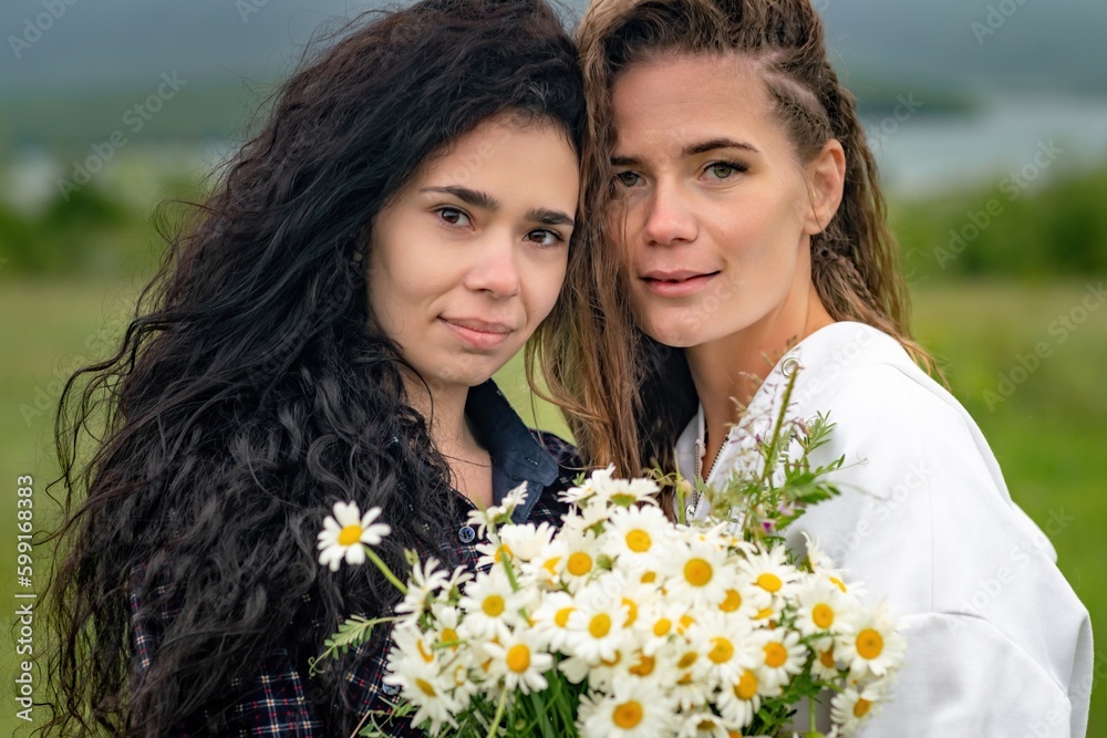 Two women enjoy nature in a field of daisies. Girlfriends hugging hold a bouquet of daisies and look at the camera. Two women enjoy nature in a field of daisies. Girlfriends hugging hold a bouquet of daisies and look at the camera.
