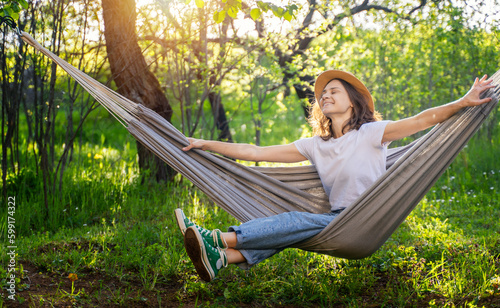 Young happy caucasian woman in a hat lying in a hammock in a green garden enjoying a summer day