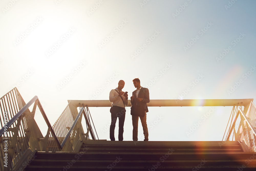 Lets set up an appointment to discuss ideas. Low angle shot of two businessmen using a cellphone on top of a staircase outside.