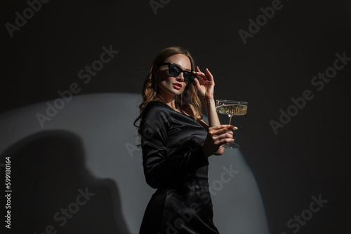 Obraz na płótnie Fashionable beautiful elegant vogue woman wears cool sunglasses in a fashion evening black dress with a glass of champagne at a party