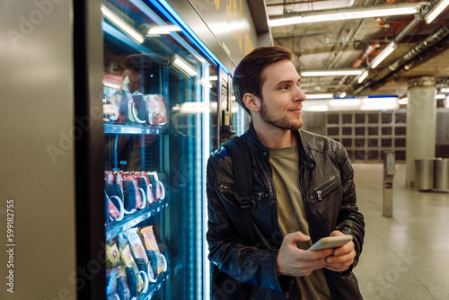 Smiling man using mobile phone while standing near snacks vending machine in subway photo