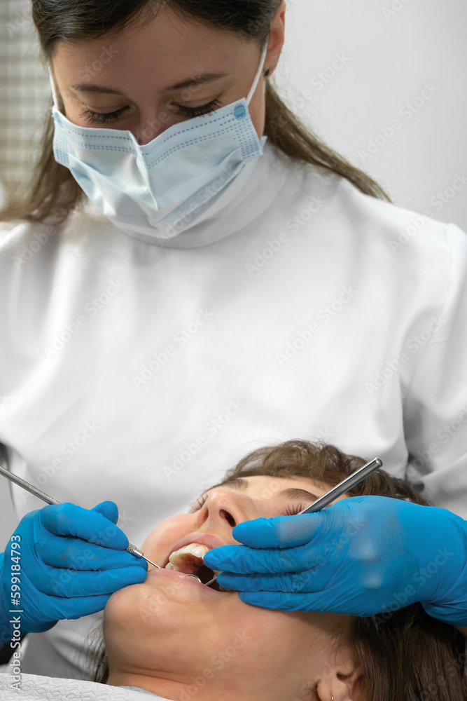 Naklejka premium Dental hygienist working with dental instruments checks patient's teeth. Teeth treatment procedure in dental clinic. Examination of teeth with dental instruments. Portrait of woman. Vertical photo