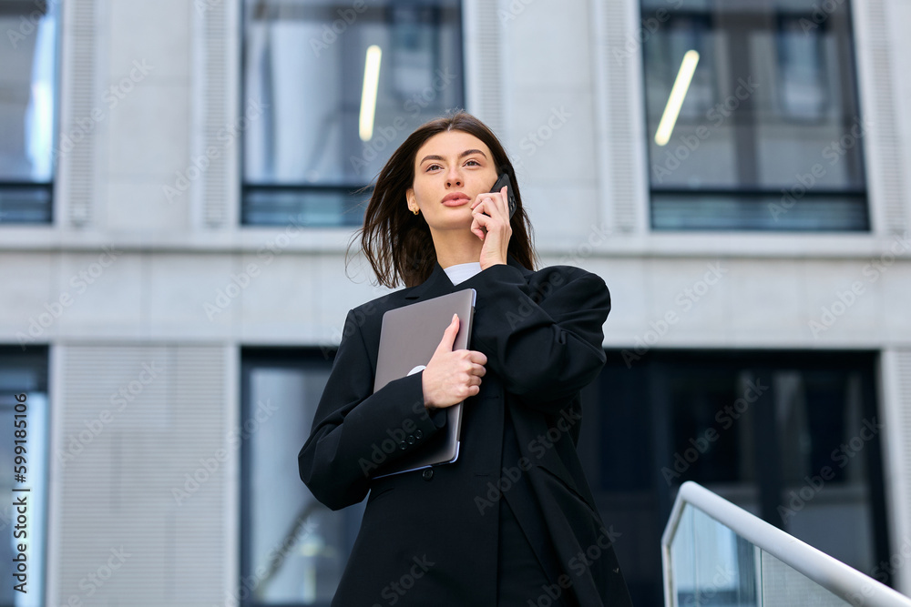 Fototapeta premium Portrait of a businesslike young woman who is talking on the phone and holding a laptop in her hands against the background of the office