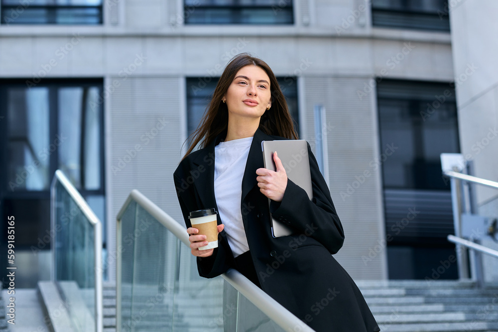 A young woman in a black suit drinks coffee and poses on the street with a laptop. Business portrait