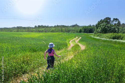 Picturesque summer landscape. A teenage girl is walking along the road through a field of young green wheat, forest and bushes in the background, against the backdrop of a blue sky.