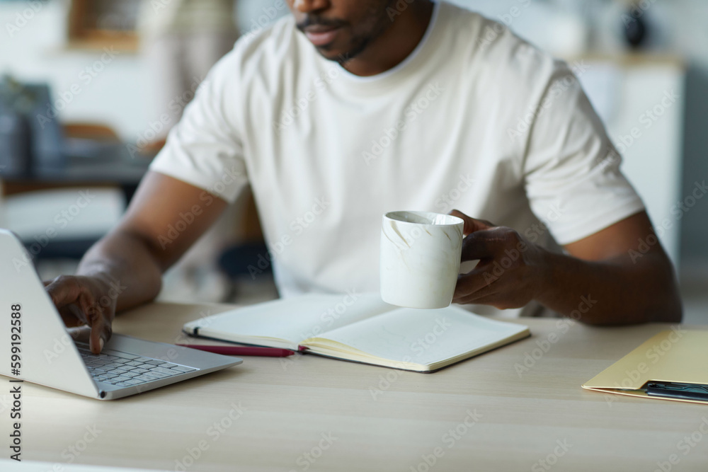 Close-up of African American man working online on laptop and drinking coffee while sitting at table in the kitchen Close-up of African American man working online on laptop and drinking coffee while sitting at table in the kitchen