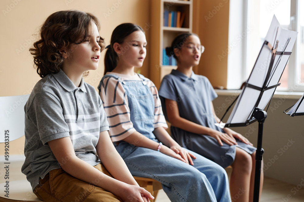Group of children sitting in front of sheet music on stand and singing together during music lesson Group of children sitting in front of sheet music on stand and singing together during music lesson