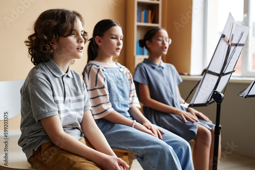Group of children sitting in front of sheet music on stand and singing together during music lesson