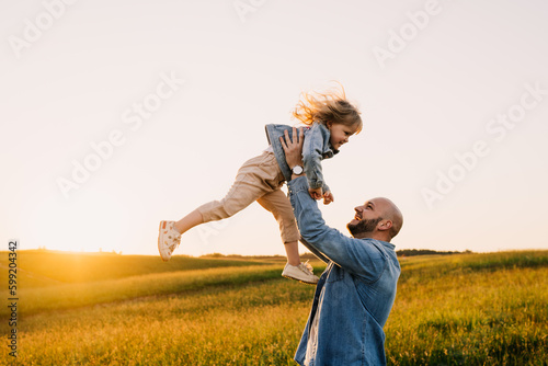 Bald caucasian young adult man throws up little daughter standing at meadow on sunset time. Happy father spends time with girl. Parenthood, relationship. Family moments, leisure, outdoor activities. #599204342