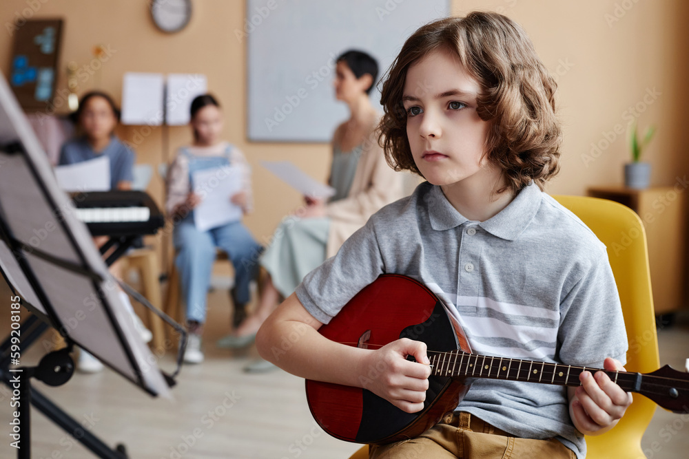 Child sitting in front of sheet music and playing musical instrument in music class with other children in background Child sitting in front of sheet music and playing musical instrument in music class with other children in background