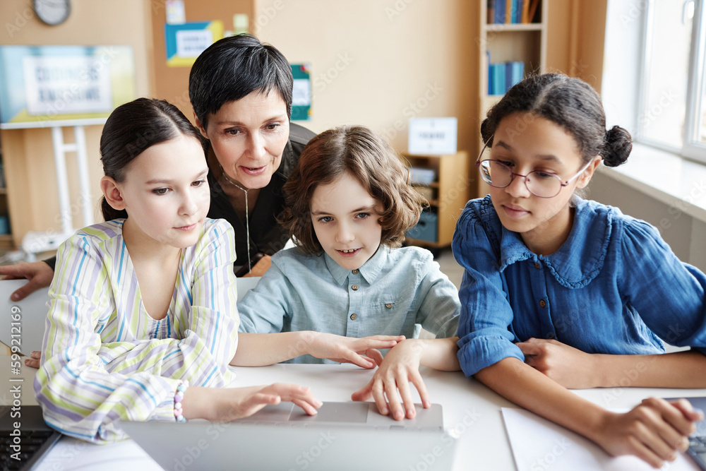 Mature teacher discussing online presentation on computer together with children during lesson in class Mature teacher discussing online presentation on computer together with children during lesson in class