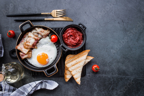Fried eggs on a stylish ceramic plate with handles with fried bacon, cherry tomatoes, tomato paste and bread toast on a dark background. Breakfast, lunch and dinner. Delicious food.