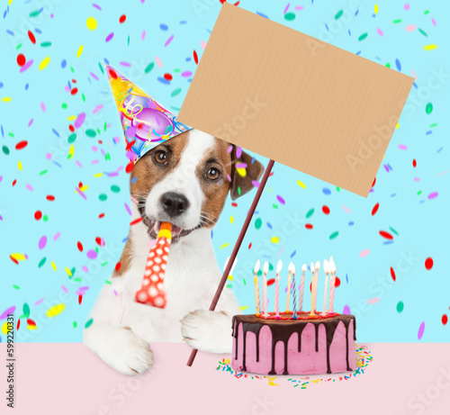 jack russell terrier puppy wearing party cap blows into party horn sits with birthday cake with lots of burning candles and shows empty placard © Ermolaev Alexandr