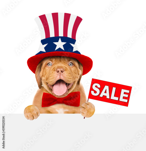 Happy Mastiff puppy wearing like Uncle Sam looks above empty white banner and shows signboard with labeled "sale". isolated on white background © Ermolaev Alexandr