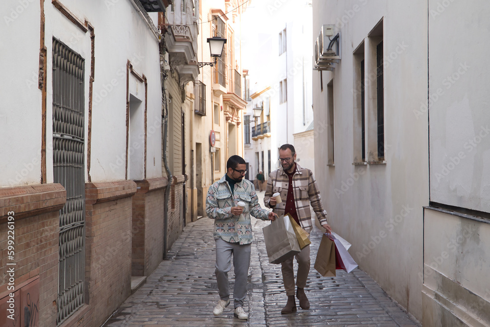 Real marriage of gay couple, walking along a lonely street, with a glass of coffee in one hand and the other full of shopping bags. Concept lgtb, lgtbiq+, couples, in love, shopping, sales.