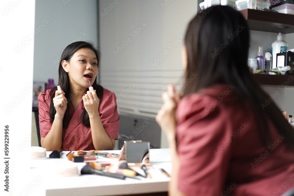 Beautiful Asian woman doing makeup and applying lipstick in front of a dressing table mirror Beautiful Asian woman doing makeup and applying lipstick in front of a dressing table mirror