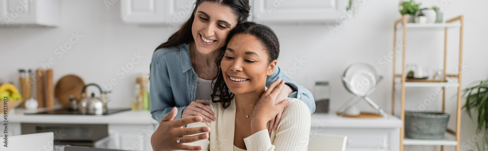 happy interracial lesbian couple hugging each other in kitchen, banner. happy interracial lesbian couple hugging each other in kitchen, banner.