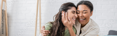 joyful multiracial woman hugging happy girlfriend in new house, banner.