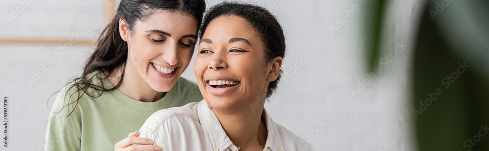 cheerful multiracial woman laughing with lesbian partner at home, banner. cheerful multiracial woman laughing with lesbian partner at home, banner.