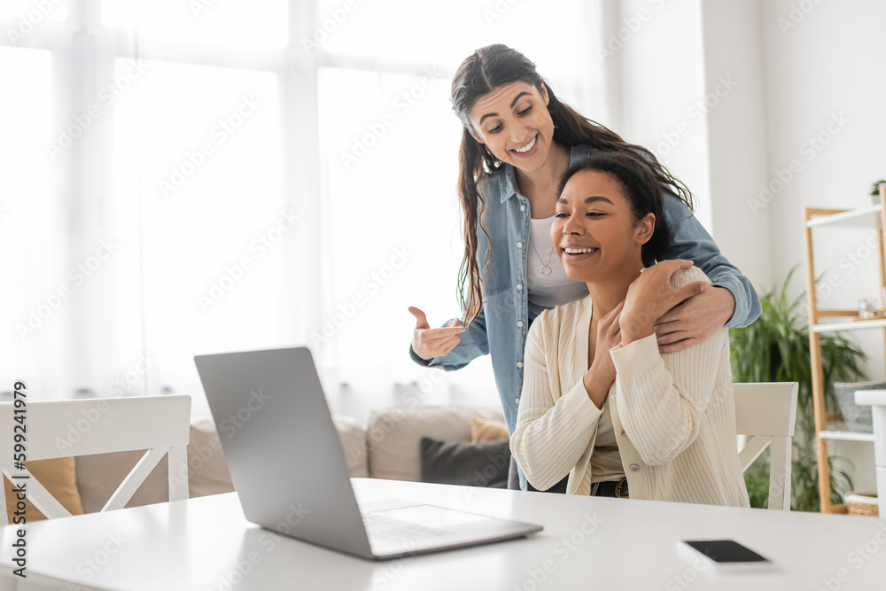 happy lesbian woman pointing at multiracial girlfriend near laptop during video call. happy lesbian woman pointing at multiracial girlfriend near laptop during video call.