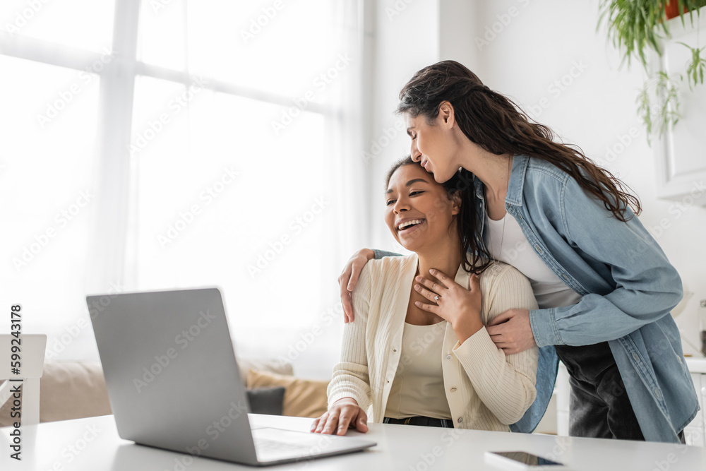 lesbian woman kissing multiracial girlfriend showing engagement ring during video call.
