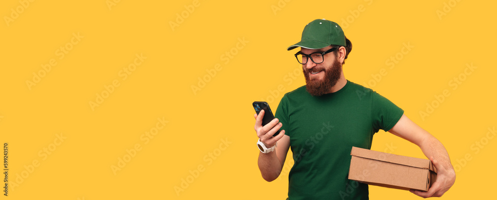 Banner shot of a man holds a box he has to deliver while looking to his phone over yellow background. - obrazy, fototapety, plakaty 