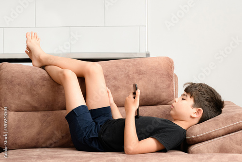The boy plays on the sofa in the living room. Living room kitchen with sofa. The child plays with gadgets. The boy spends his summer vacation playing games on his phone.