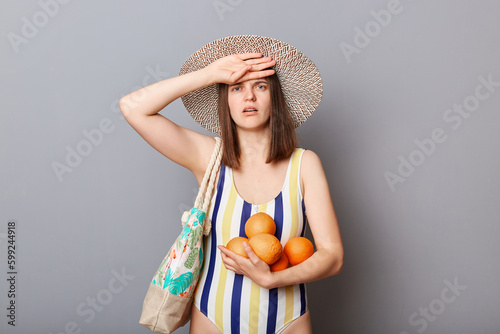 Sick unhealthy tired woman wearing striped swimsuit and straw hat  isolated gray background having headache expressing sorrow has sunstroke feeling unwell.