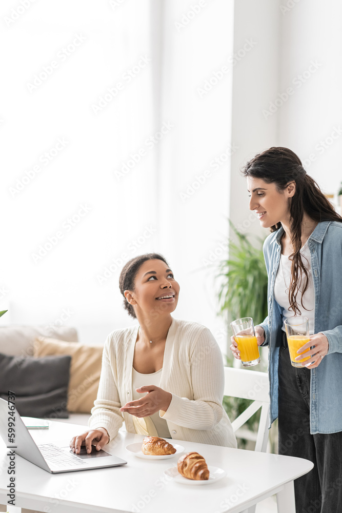 cheerful lesbian woman holding glasses with orange juice near multiracial partner working on laptop.