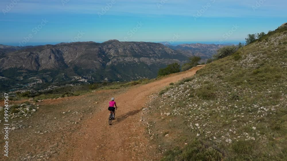 Gravel cycling adventure with beautiful mountain and sea view.Woman athlete wearing cycling kit,pink jersey and white helmet.Outdoor training.Spain