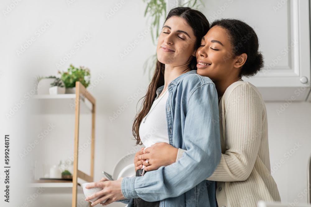 cheerful multiracial woman with engagement ring on finger hugging girlfriend holding cup of coffee. cheerful multiracial woman with engagement ring on finger hugging girlfriend holding cup of coffee.