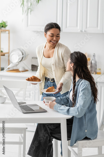 multiracial woman holding plates with baked croissants near happy girlfriend working on laptop from home. © LIGHTFIELD STUDIOS